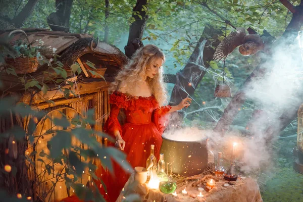 Mooie jonge dame met blond krullend haar boven grote magische ketel met rook en flesjes met vloeistoffen, bos nimf in lang lichte rode jurk met losse mouwen bereidt potion in de buurt van houten huis — Stockfoto