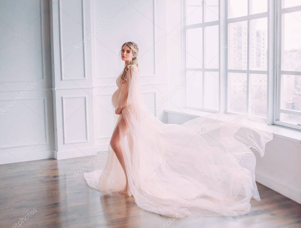 cute young slim lady with tummy in spacious room with white walls and large windows, pregnant girl with blond hair twisted in a long pink gentle lace peignoir with flying train posing for camera