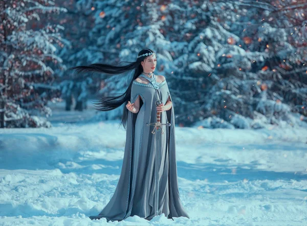 Karlı ormanda büyüleyici çekici Bayan, siyah uzun uçan saçlı militan elf Prensesi, kış aylarında ateş kıvılcımları, yaratıcı soğuk mavi renkler kılıç, gevşek gri sıcak elbise ve yağmurluk tutar — Stok fotoğraf
