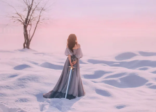 Mysterieuze dame uit middeleeuwen met donker haar in zachte grijze blauwe jurk in besneeuwde woestijn met open rug en schouders, scherp zwaard achter, model staat terug met geen gezicht, creatieve pastelkleuren — Stockfoto