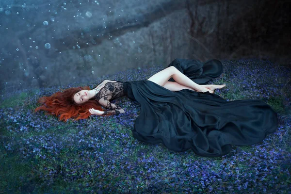 Dziewczyna z czerwonymi włosami leżącą na trawie w ciemnym lesie, czarna królowa zagubiona w bitwie, urocza dama w długiej czarnej królewskiej sukni z koronką na otwartej klatce piersiowej, seksowna gorąca nimfa z długimi gołymi nogami w najważniejszych księżycowych — Zdjęcie stockowe