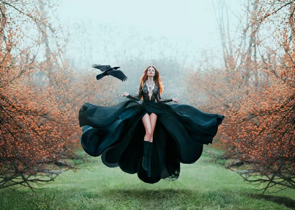 Girl with bright red hair levitates above ground, powerful sorceress, forest goddess in black flying dress with lace on open chest and bare long legs, lady in flowering orange garden and hand raven Stock Photo