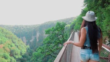 young woman in denim shorts with her back to camera, with long straight black hair and hat on head, sporty lady touches iron railing, looking down on Okatse canyon, amazing nature of Georgia