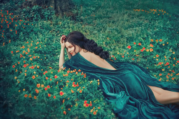 Charming nymph lying on green grass and bright small flowers, girl in a long emerald velor dress with open bare back is resting in a clearing, forest fairy with dark hair observes life of forest.