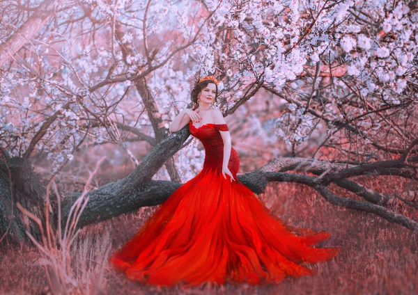 Encantadora ninfa senta-se na árvore caída na floresta da primavera, senhora em lindo vestido longo vermelho escarlate com ombros nus, sereia se torna humana, princesa de conto de fadas caminha no jardim sakura, cores criativas — Fotografia de Stock