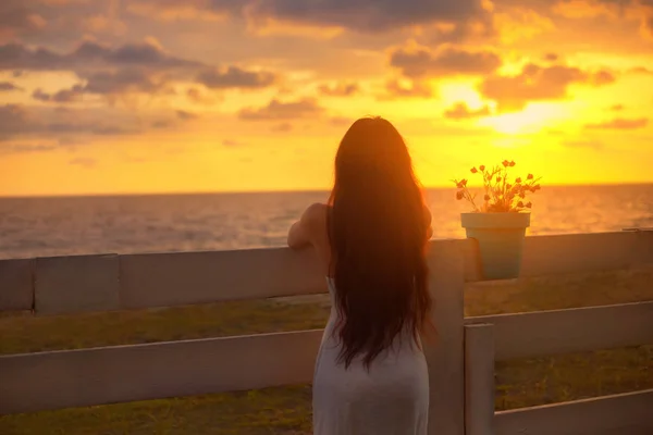 Fille aux cheveux bruns avec une silhouette chic mettre ses mains sur une clôture avec un pot de fleurs, une dame en robe blanche, regarde coucher de soleil rouge vif et jaune dans la mer avec un ciel nuageux. photo sans visage de dos — Photo