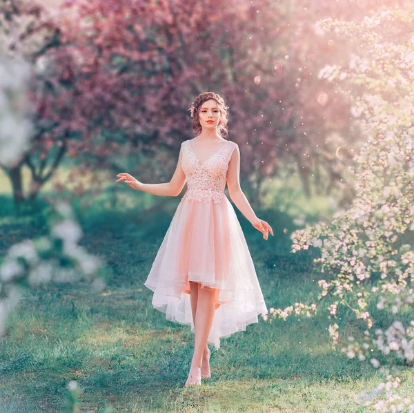 Geheimnisvolle Dame mit dunklem gerafftem Haar im hellen zarten Pfirsichkleid spaziert durch den blühenden Garten, die Frühlingsfee berührt ihre Habseligkeiten, die Märchenelfe wie eine Puppe barfuß auf dem Gras — Stockfoto