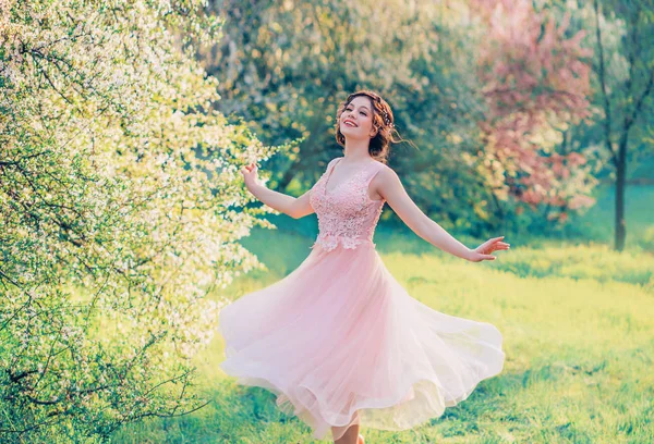 Chica feliz en vuelo corto suave vestido rosa se ríe alegremente, muñeca princesa gira en brillante jardín de primavera amarillo con árboles florecientes, emociones positivas, movimiento en la foto con colores creativos —  Fotos de Stock