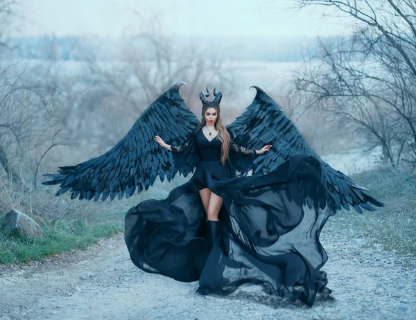 迷人的华丽黑暗女神控制风，气流波下交和长火车浅黑色礼服与宽蕾丝袖子，女士与锋利的角和黑色羽毛翅膀准备飞向天空 — 图库照片