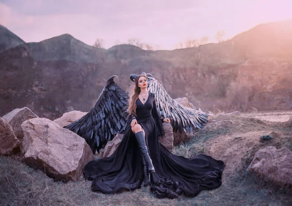 Revived gargoyle, queen of night watching sunrise, girl in long light black dress with black feather wings sits on rocks with open leg in high boots, mysterious mythical creature, creative art photo — Stock Photo, Image