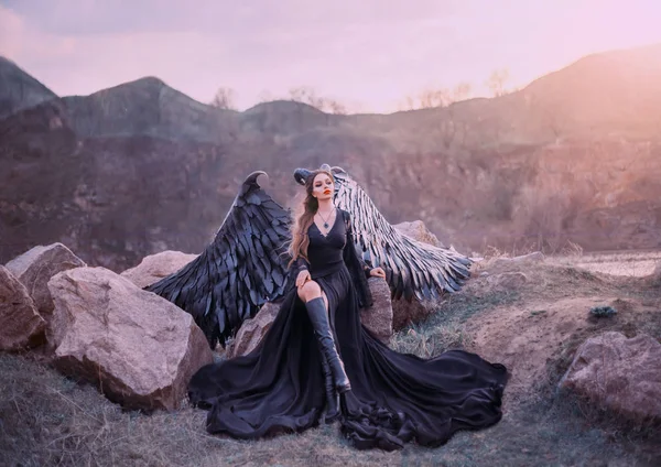 Gárgola revivido, reina de la noche viendo la salida del sol, chica en vestido largo negro claro con alas de plumas negras se sienta en las rocas con la pierna abierta en botas altas, misteriosa criatura mítica, arte creativo foto — Foto de Stock