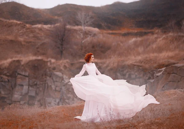 Belleza pelirroja en un vestido vintage blanco, se encuentra en la cima de una colina, fondo de las montañas rocosas. Lujoso atuendo con un largo tren revoloteando en el viento. Tejidos de seda voladores ligeros. colores cálidos —  Fotos de Stock