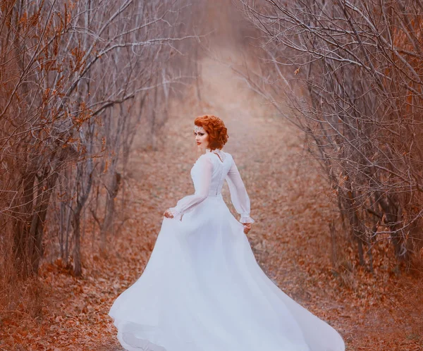Рыжая принцесса в белом винтажном платье бежит в туннеле с деревьев и оглядывается назад. Роскошный наряд с длинным трепетом поезда на ветру. Спящая природа, золотая осень, теплые цвета — стоковое фото