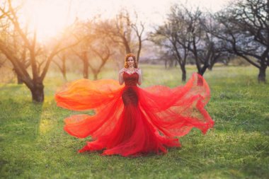 Kırmızı elbiseli doğu kraliçesi rüzgarda çırpınan uzun bir tren ile bir denizkızı bir siluet. Bir taç ile zarif saç modeli. Altın ve yakut ile lüks kolye. Bahçedeki kadın