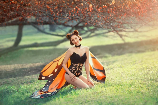 Χαριτωμένο κοκκινομάλλα κορίτσι κάθεται σε ζουμερό φρέσκο γρασίδι, δάσος νεράιδα με χαμόγελο, ελκυστική κυρία στην εικόνα της πορτοκαλί και κίτρινη πεταλούδα, σέξι κορίτσι με μαύρο κορμάκι με δημιουργικό μοτίβο και ελαφριά φτερά — Φωτογραφία Αρχείου