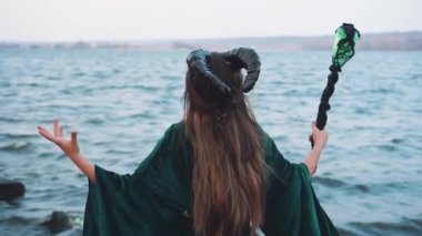 nehir kenarında elinde sihirli bant ile altın kemer ile uzun yeşil elbise li baş siyah büyük boynuzları ile bayan, zümrüt manto içinde ağartılmış saçlı cadı kader hakkında cennet sorar, yüz olmadan arkadan