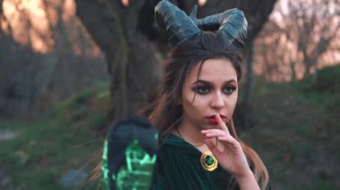 başında büyük keskin siyah boynuzları ile gizemli efsanevi yaratık kırmızı parlak dudaklar, elinde sihirli sopa ile uzun yeşil zümrüt manto kız parmak tutar. cadı sırrını saklamak istiyor