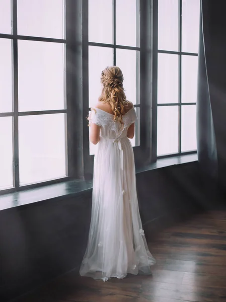 Femme mystérieuse loking à de grandes fenêtres lumineuses, princesse enchantée se transforme en beau cygne, tissage créatif pour les cheveux blonds, idée pour l'image du bal de promo pour les jeunes filles, pas de visage avec son dos à la caméra — Photo