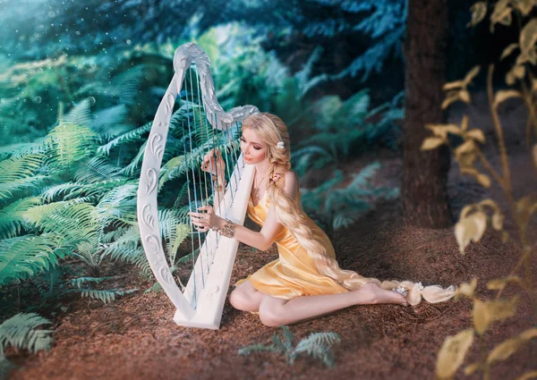 Fabulous forest elf sits under tree and plays on white harp, girl with long blond hair braided in long yellow dress, summer goddess rests and sings to the sound of a magical musical instrument — Stock Photo, Image