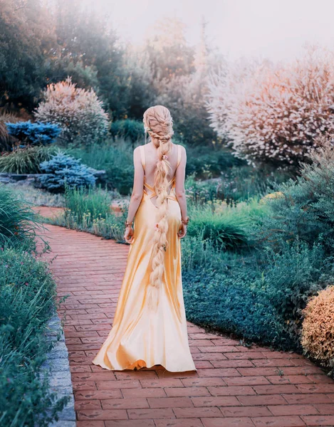 Misteriosa signora in chic giallo costoso vestito di lusso in magnifico giardino, principessa misteriosa con lunghi capelli biondi intrecciati in piedi capelli con le spalle alla macchina fotografica, fata mistica della natura, senza volto — Foto Stock