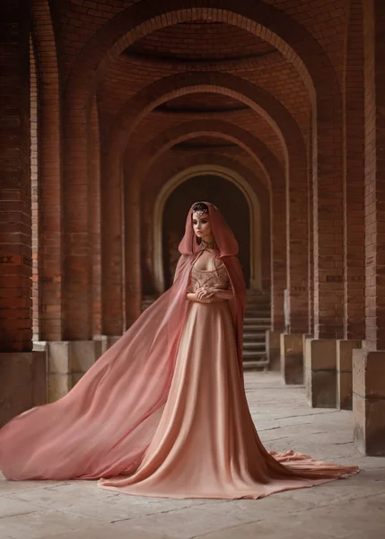 Een luxe dame in een lange roze jurk in een Road Coat met een capuchon loopt langs de oude kasteel gang. De Elven koningin. De stof van de trein vliegt, zwaaiend. Kunst samenzweringen en intriges. — Stockfoto