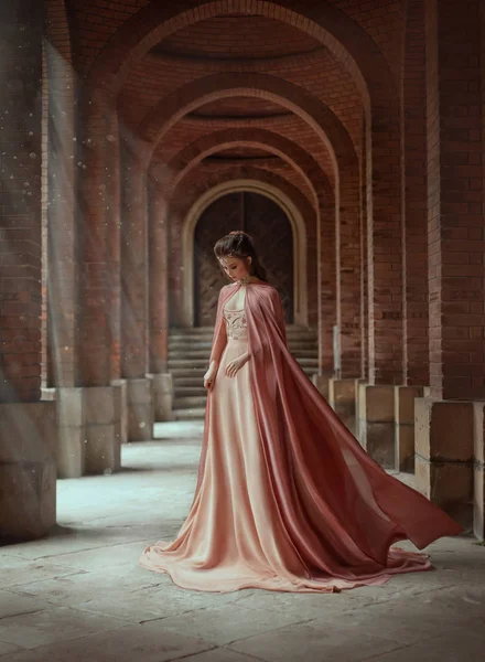 Sad princess in a vintage royal nude powdery color dress and in a cloak that flies in the wind. The magical Rays of the sun and hope pour on a woman through the columns. Long dark hair with a tiara. — Stock Photo, Image