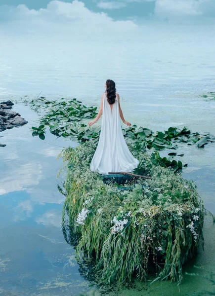 Una donna misteriosa in un lungo abito bianco galleggia su una barca per incontrare il cielo blu e le nuvole. Arredamento con erba, rami e fiori. Matrimonio creativo insolito. Sparare dalla schiena senza una faccia — Foto Stock