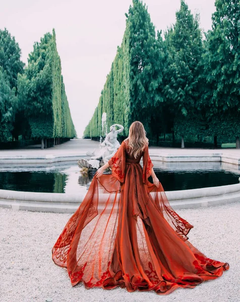 Misteriosa reina en un peignoir largo transparente rojo con un tren. Atractiva mujer rubia con su cabello camina en un lujoso jardín con una fuente. Fondo de árboles verdes de corte alto, como una pared . — Foto de Stock