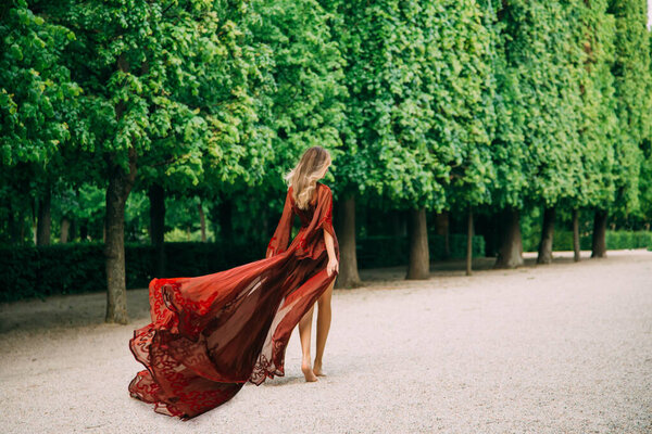 Tall woman in a red silk dress with a long train that flutters in the wind. Medieval fashion of luxurious wide-sleeve peignoirs. Summer day. Happy valentines day. The girl goes to meet her beloved.
