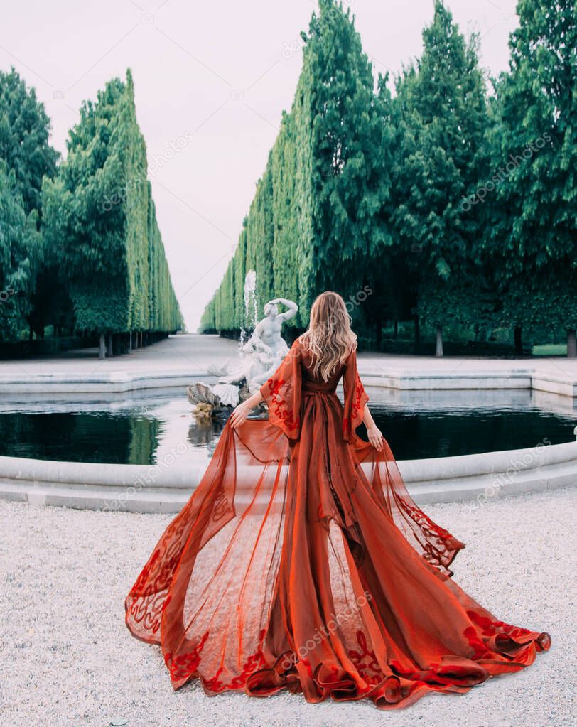 Mysterious queen in a red transparent long peignoir with a train. Attractive blond woman with her hair walks in a luxurious garden with a fountain. Background of tall cut green trees, like a wall.