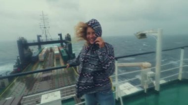 Bir kadın, fırtınada bir kargo gemisinin güvertesinde duruyor. Şiddetli rüzgar fırtınaları, düşmemek için tırabzanları sıkıca tutuyor. Denizde kötü hava. Kapüşonlu kız gülümsüyor, olumlu düşünüyor..