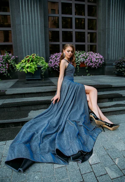Basamaklarda oturan güzel genç kadın model modern şehir binası, çiçek açan petunya çiçekleri. Şık, uzun, gri, mavi, zarif balo elbisesi. Seksi çıplak bacaklar, siyah ayakkabılar. Eski moda saç stili — Stok fotoğraf