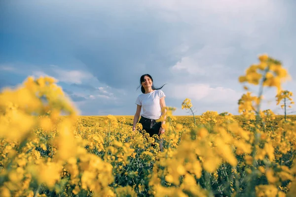Jovem alegre mulher feliz caminha no campo de colza florescendo amarelo. Nuvens escuras céu azul. Morena menina divertida sorrindo, alegria desfrutar de beleza primavera aroma flores verão natureza. Conceito livre de alergia — Fotografia de Stock