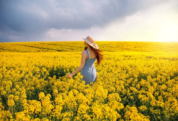 Jovem mulher caminhando campo de floração, virou-se, toque suavemente flores amarelas. Menina morena gosta de aromas de primavera, harmonia. Fundo nuvens escuras céu azul. Longo cabelo vacilante vento. Chapéu de vestido de ganga — Fotografia de Stock