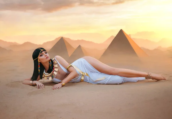 Egypt Style Rich Luxury Woman. Sexy beautiful girl goddess Queen Cleopatra lies on yellow sand desert pyramids. Art ancient pharaoh costume white dress gold accessories Black hair wig Egyptian makeup — Stock Photo, Image