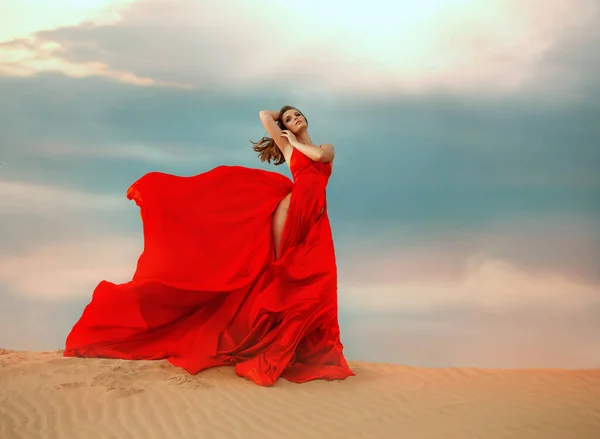 Giovane donna sexy regina glamour stand. Bellissime dune di sabbia del deserto drammatiche nuvole di cielo. Abito lungo in raso di seta rosso elegante, gonna a spacco sulle gambe, mosca svolazzante in movimento vento. Ragazza modella posa — Foto Stock