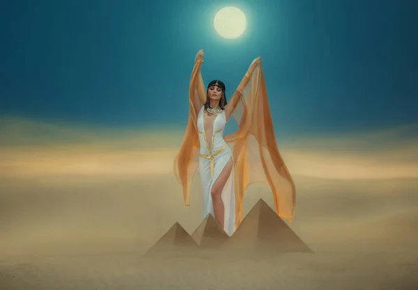 Konstverk Fantasy egyptiska skönhet gudinnan Cleopatra höjer händerna till blå natthimmel, bakgrund pyramid gul sand sanddyn ljus måne. Orange gyllene mantel vit sexig klänning. Svart hårdrottning Nefertiti — Stockfoto