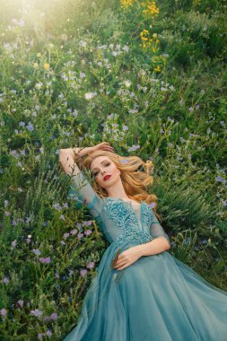 Genç ve güzel bir kadın yeşil çimlerin üzerinde yatıyor. Mor sarı çiçekler. Mavi lüks gece klasik tül elbisesi. Kız prenses yaz doğasını seviyor. Sarışın, uzun dalgalı saçlı, kırmızı dudaklı, romantik yüzlü.