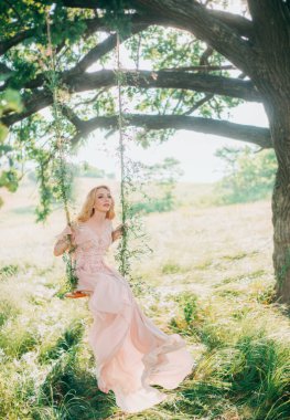 Sihirli salıncakta oturan güzel kadın perisi. Uzun bej şeftali rengi ipek, eski moda elbise. Gelin Prenses sarı saç. Güzel sanatsal düğün fotoğrafı. Sonbahar sisli arka plan doğa yeşil çimen yaz ağaçları