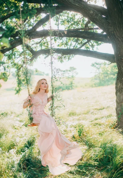 Mooie vrouw nimf zitten op magische swing. Lange beige perzik zijde vintage mode jurk. Bruid Prinses blond haar. Mooie kunst trouwfoto. Herfst mistige achtergrond natuur groen gras zomer bomen — Stockfoto