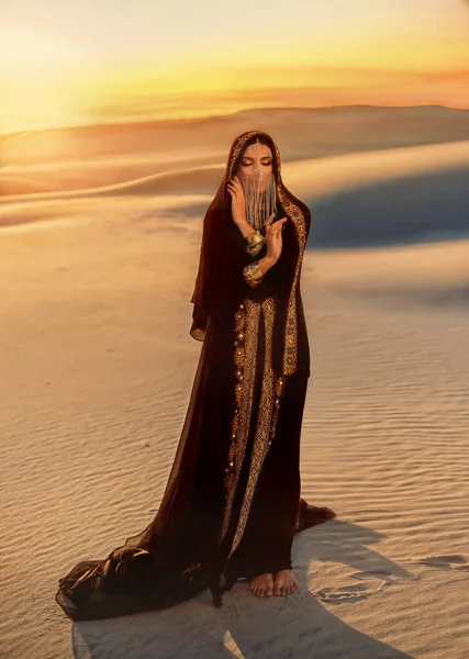 Mulher árabe misteriosa bonita em vestido preto muçulmano abaya. cabeça é torcida com seda lenço cadeia esconde rosto. Fantasia menina caminha no deserto, arte fundo areia ouro por do sol. Rainha Concubina — Fotografia de Stock
