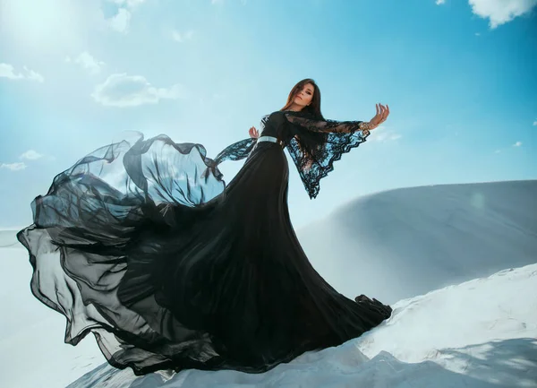 Όμορφη μυστηριώδης Άραβα βασίλισσα με μαύρα ρούχα. Κοριτσίστικο μοντέλο που ποζάρει, χορεύει σε μακρύ, μεταξωτό φόρεμα, υφασμάτινες μύγες σε κίνηση ανέμου. Πριγκίπισσα περπατά στην έρημο, φόντο λευκή άμμο μπλε ουρανό — Φωτογραφία Αρχείου