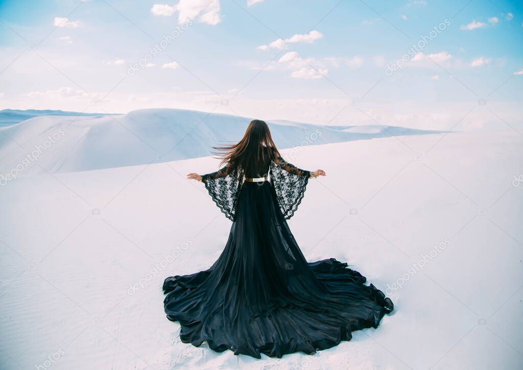 Rear view Silhouette of Beauty woman. Queen in black clothes stands in desert. Girl fashion model. long, silk dress with train. Back of luxury glamorous elegant goddess. background white sand blue sky