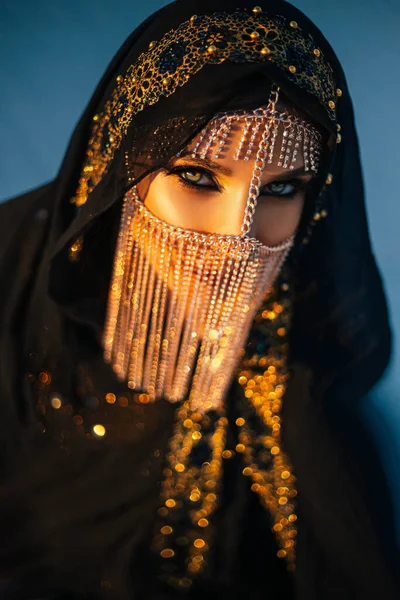 Portrait of young beautiful oriental mysterious girl. The woman looks into the camera. The face is hidden by a golden veil. Luxurious evening make-up, smoky eyes. The head is covered with black scarf — Stock Photo, Image