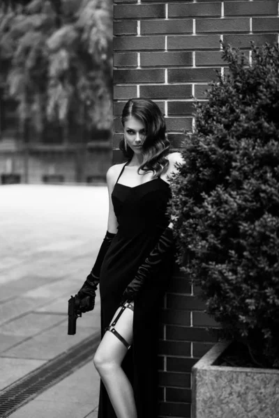 Krásná mladá dívka v elegantních šatech. Žena držící v rukou zbraň. Módní model v podobě nebezpečného sexy špióna. Černobílá fotografie. Retro dáma, retro účes večerní make-up — Stock fotografie