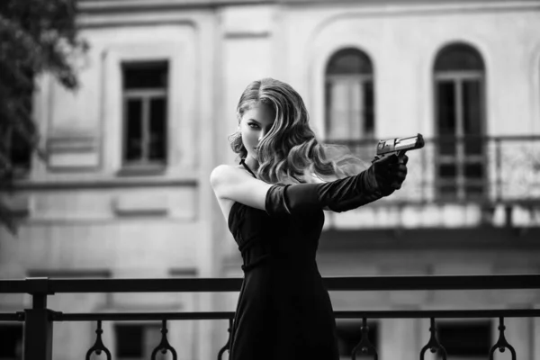 Hermosa joven con un vestido elegante. Mujer sosteniendo un arma en sus manos. Modelo de moda en forma de un peligroso espía sexy. Fotografía en blanco y negro. Señora retro, peinado vintage maquillaje de noche —  Fotos de Stock