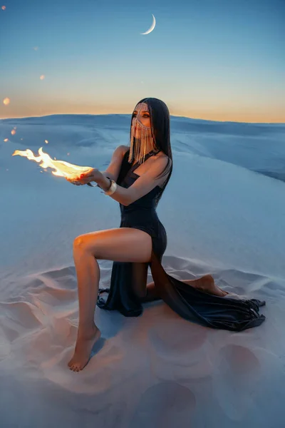 Een mysterieuze vrouw zit op het zand en houdt een vuur in handen. Fantasie, kunstfoto. Het meisje verbergt haar gezicht achter een gouden sluier. Een vrouw in de gedaante van een tovenaar. Woestijnnacht achtergrond. Creatieve jurk met spleten — Stockfoto