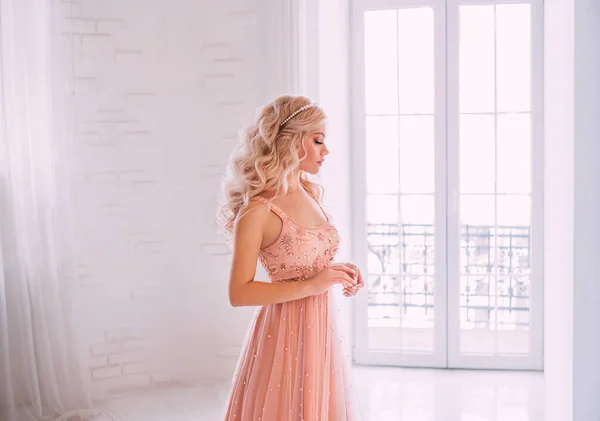 Retrato de uma jovem mulher bonita. Olhos fechados. Cosmético natural. Pearl Tiara cabelo ondulado longo loiro. Bege cor pêssego vestido de luxo bordado. Interior da sala branca de pano de fundo, janela clássica alta — Fotografia de Stock