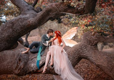 Birbirine aşık genç bir çift. Bir adam ve bir kadın kocaman bir ağaçta sarılıyorlar. Temalı yaratıcı düğün parlak fantezi fotoğrafçılığı. Parlak altın kanatlı, uzun pembe elbiseli peri kadın. Elf kostümü giymiş bir adam.
