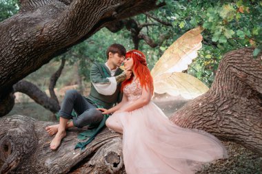 Birbirine aşık genç bir çift. Bir adam ve bir kadın kocaman bir ağaçta sarılıyorlar. Temalı yaratıcı düğün parlak fantezi fotoğrafçılığı. Parlak altın kanatlı, uzun pembe elbiseli peri kadın. Elf kostümü giymiş bir adam.